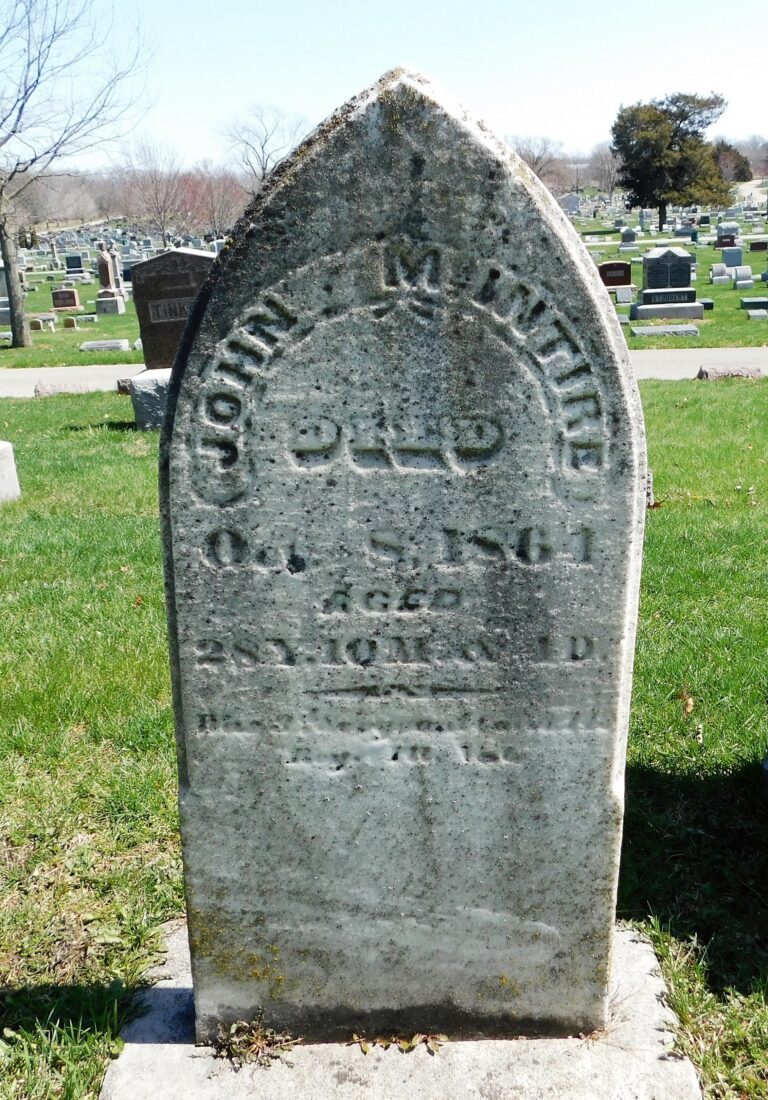 Weathered arched tombstone in a grassy cemetery with many headstones in the background, inscription now worn away.