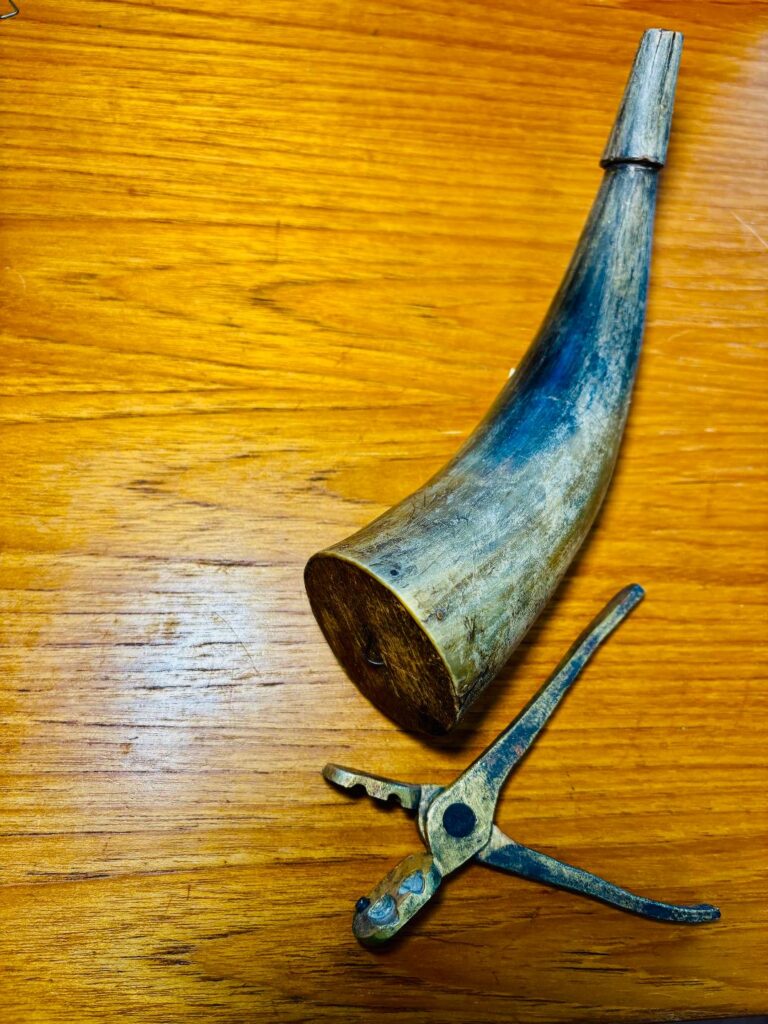 Old curved horn (likely a powder horn) on a wooden table, with a small rusty metal tool nearby.