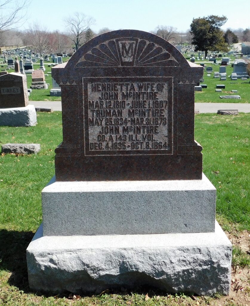 Granite grave marker with a rounded top and sunburst carving, displaying an engraved inscription on the front, set among many gravestones on a green cemetery lawn.
