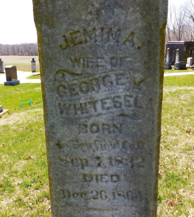 Weathered grave marker in a cemetery with grass around it and inscriptions barely readable due to weathering.