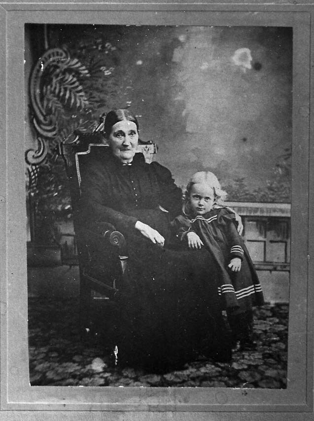 Older woman in a dark dress sits in an ornate chair with a young girl in a sailor-style dress beside her, in a studio backdrop.