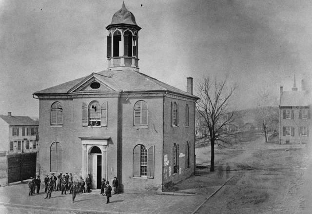 Black-and-white image of a small town church with a cupola, crowds gathered at its entrance on a quiet street.