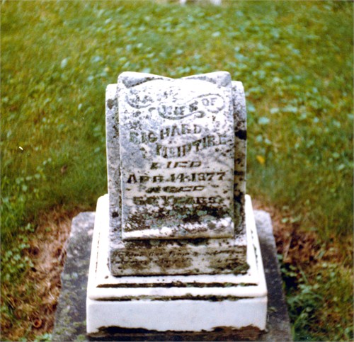 Weathered tombstone in a grassy cemetery, inscription faded and difficult to read.
