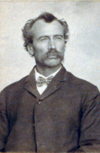 Portrait of a 19th-century man with a mustache and wavy hair, wearing a dark suit and bow tie, looking off to the side