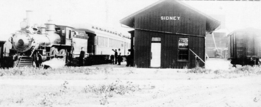 Steam locomotive with passenger cars beside a dark SIDNEY station building; several people stand nearby in a vintage rail yard.