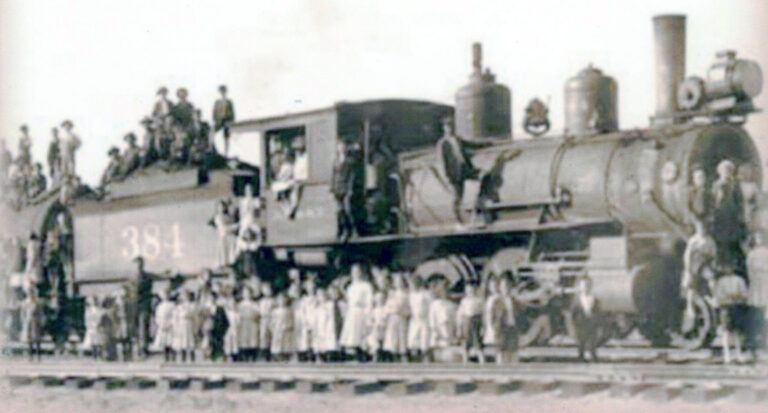 Vintage steam locomotive with a large crowd gathered on and beside it along the railroad tracks, resembling a parade or public event.