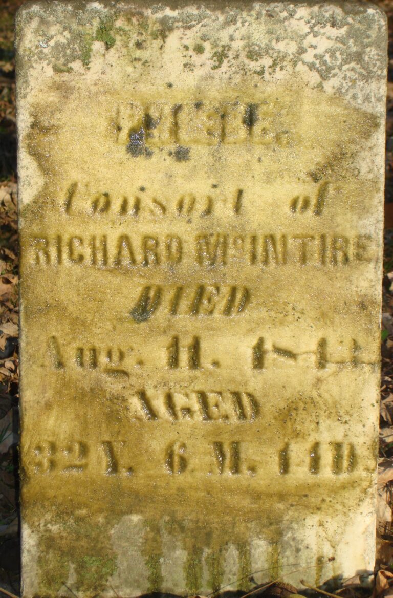 Weathered, moss-covered tombstone with eroded inscription; text is mostly unreadable due to age and staining.
