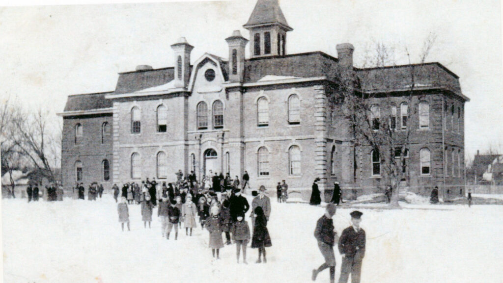 Brick schoolhouse or town hall in winter, with a crowd gathered outside in snowfall.