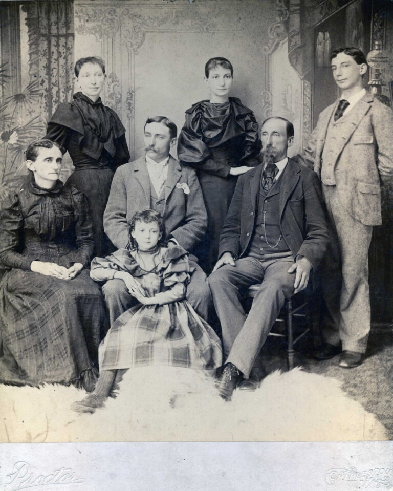 Studio portrait of a multi-generational family from the early 20th century: two men in suits, three women in high-neck dresses, and a young child seated at the front, posed against a decorative backdrop.
